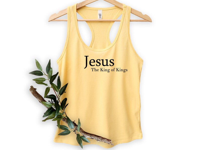 May include: A light yellow tank top with a racerback design. The text "Jesus The King of Kings" is printed in black on the front. The tank top is hanging on a wooden hanger. A branch with green leaves is placed in front of the tank top.