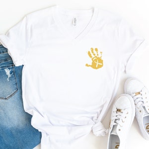 May include: White v-neck t-shirt with a gold handprint and a white ribbon on the front. The shirt is paired with blue jeans and white sneakers.
