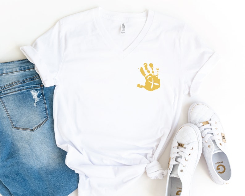 May include: White v-neck t-shirt with a gold handprint and a white ribbon on the front. The shirt is paired with blue jeans and white sneakers.