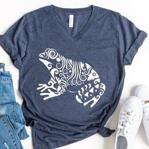 May include: A dark heather blue V-neck t-shirt featuring a large white graphic of a frog decorated with intricate swirling patterns and lines. The shirt has short sleeves and a visible brand tag at the inner neckline, presented as a flat lay.