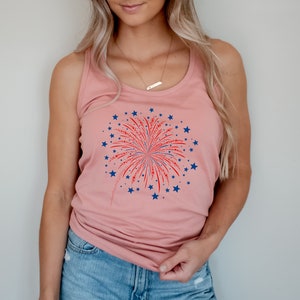 Puede incluir: Una camiseta de tirantes rosa con un gr&aacute;fico de fuegos artificiales rojo, blanco y azul. Los fuegos artificiales est&aacute;n rodeados de estrellas azules.