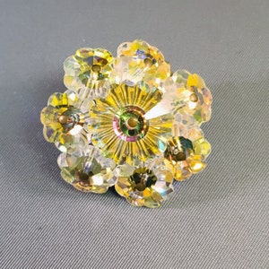 Può includere: Una spilla a forma di fiore scintillante con cristalli iridescenti. La spilla presenta un design centrale dorato circondato da sei petali di cristallo. I cristalli riflettono un arcobaleno di colori, creando un effetto abbagliante. La spilla è probabilmente in vetro o cristallo.