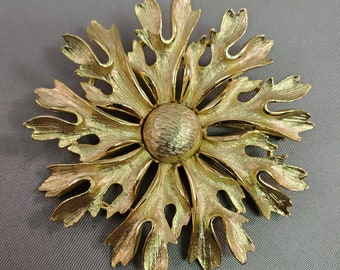 May include: A gold-toned metal brooch in a floral design. The brooch features a central textured circle surrounded by stylized, leaf-like petals. The petals have a textured finish and are arranged in a symmetrical pattern.