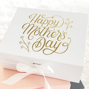 May include: A white gift box with a gold inscription that reads "Happy Mother's Day" with a floral design. The box is tied with a white ribbon and is stacked on top of a pink gift box with a pink ribbon. The pink box is on top of a black gift box.