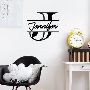 May include: Black and white vinyl wall decal with the letter J and the name Jennifer written below it. The decal is on a white wall with a clock and a chair in the background.
