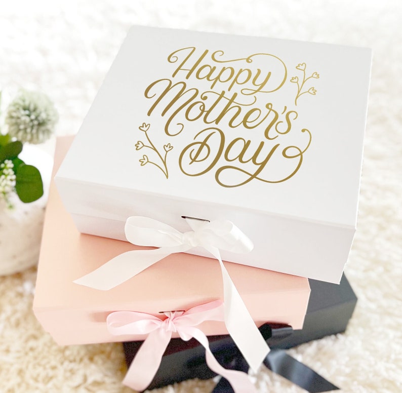 May include: A white gift box with a gold inscription that reads "Happy Mother's Day" with a floral design. The box is tied with a white ribbon and is stacked on top of a pink gift box with a pink ribbon. The pink box is on top of a black gift box.