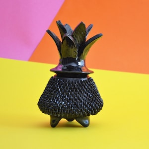 May include: Black ceramic pineapple shaped vase with a textured surface and three legs. The vase is on a yellow, pink, and orange background.