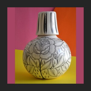 Puede incluir: Jarra de cerámica blanca con un diseño floral en blanco y negro. La jarra tiene un cuerpo redondo y una boca ancha. El diseño floral presenta una rosa grande con hojas y tallos.