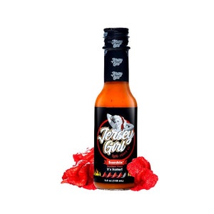 Puede incluir: Una botella de salsa picante Jersey Girl de 148 ml con una etiqueta roja y negra. La etiqueta presenta a una mujer con un vestido rojo y el texto "Scorchin' Sodium Free It's Hotter!" La botella está rodeada de chiles rojos.
