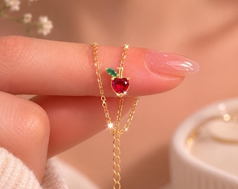 May include: A gold necklace featuring a pendant shaped like a red heart-shaped apple with a green leaf. The necklace is held between fingers, showcasing the delicate chain and pendant design. The apple pendant is approximately 0.5 inches in height.