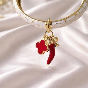 Può includere: Un bracciale rigido color oro con inserti in smalto bianco e un motivo circolare ripetuto. Tre ciondoli pendono dal centro: un quadrifoglio rosso, un peperoncino rosso e un ciondolo a forma di lettera R color oro con finitura metallica.