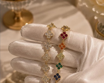 May include: Two gold-tone bracelets featuring a series of four-leaf clover motifs. One bracelet is adorned with clear, sparkling stones, while the other features a vibrant rainbow sequence of colored stones including red, orange, yellow, green, blue, and purple.
