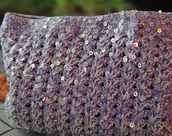 May include: A lavender-colored, crocheted clutch bag with silver sequins. The bag has a textured, woven pattern and a rectangular shape. The sequins are scattered across the surface, adding a touch of sparkle. The background is blurred, with hints of green.