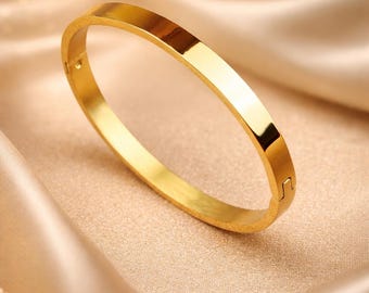 May include: A gold-colored bangle bracelet with a rectangular design. The bracelet is made of a shiny, reflective metal and is resting on a satin fabric. The bracelet has a clasp closure. Jewelry.