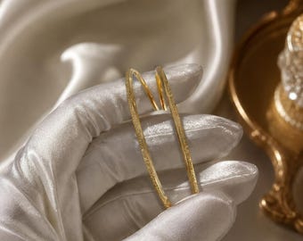 May include: A pair of gold-colored hoop earrings. The earrings are held by a gloved hand against a backdrop of white satin fabric. The earrings have a textured surface and a simple, elegant design.