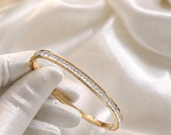 May include: A gold-colored bangle bracelet with a row of small, clear gemstones set along the top. The bracelet is shown against a white satin background. The bracelet is a piece of jewelry.