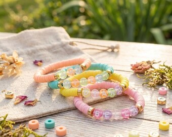 May include: A collection of colorful beaded bracelets with gold accents, spelling out names. The bracelets are in shades of pink, yellow, turquoise, and peach. The bracelets are on a wooden surface with scattered beads and dried flowers.