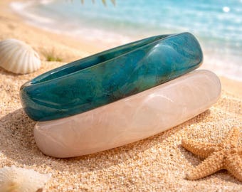 May include: Two smooth, colorful bangles rest on a sandy beach with seashells and a starfish. The top bangle is teal, and the bottom is a light pink. Palm fronds and the ocean are in the background.