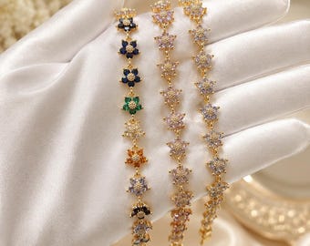 May include: Three gold-tone chain bracelets featuring a series of flower-shaped charms. The left bracelet displays multicolored gemstones including blue, green, orange, and purple. The two right bracelets feature light purple and clear crystal stones.
