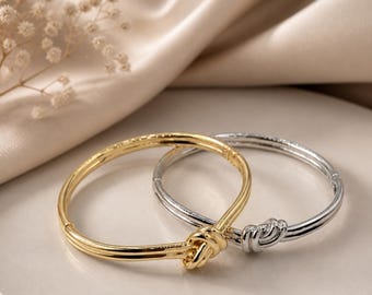 May include: Two bangle bracelets, one gold-toned and one silver-toned, each featuring a knot design at the center. The bracelets are displayed on a white plate against a soft, neutral background. The gold bracelet has a ribbed texture.