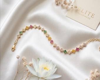 May include: A gold-toned chain bracelet featuring a series of small, four-petaled flower charms. Each flower is encrusted with tiny, multicolored gemstones in shades of pink, yellow, green, and clear white. The bracelet includes a lobster claw clasp.