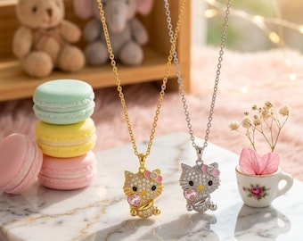 May include: Two necklaces with pendant charms in the shape of a cat. One necklace is gold-toned, the other is silver-toned. The cat pendants are encrusted with small, sparkling stones and have pink bows and heart accents. The necklaces are displayed on a marble surface.