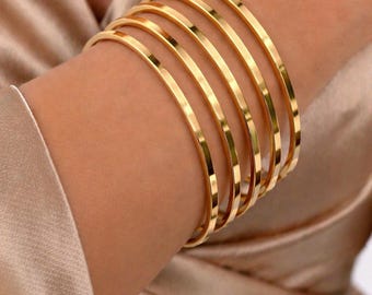 May include: A collection of gold-colored bangles stacked on a wrist. The bangles are thin and have a smooth, reflective surface. The arm is adorned with multiple bangles, creating a layered effect. The background is a soft, satin fabric.