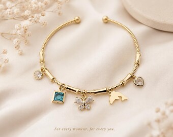 May include: A gold-toned open bangle bracelet featuring a textured chain design with five dangling charms: a clear round stone, a square blue stone, a crystal-encrusted butterfly, a dolphin silhouette, and a heart-shaped stone. Text reads: For every moment, for every you.