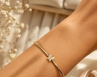 May include: A gold-colored bracelet with a butterfly charm. The butterfly is encrusted with small, clear gemstones. The bracelet is thin and delicate, with a simple design. Other bracelets are visible in the background.
