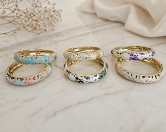 May include: Six gold-toned bangles with colorful floral enamel designs. The bangles feature shades of blue, pink, green, white, and purple. The jewelry is displayed on a marble surface, with a soft, neutral-colored fabric in the background.