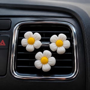 Könnte beinhalten: Drei weiße, margeritenförmige Auto-Lufterfrischer mit gelben Zentren sind an einer schwarzen Lüftungsöffnung befestigt. Die Lufterfrischer sind in einer Reihe angeordnet, mit der Lüftungsöffnung und dem Armaturenbrett im Hintergrund.
