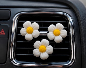 May include: Three white daisy-shaped car air fresheners with yellow centers are attached to a black car vent. The air fresheners are in a row, with the car vent and dashboard in the background.