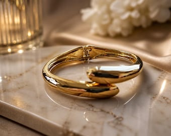 May include: A gold-colored hinged bangle bracelet with a smooth, curved design. The bracelet is highly reflective, catching the light and creating a bright, polished appearance. The jewelry is displayed on a marble surface.