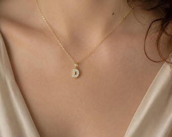 May include: A gold-toned chain necklace featuring a small, rectangular pendant shaped like the letter D. The surface of the letter D is encrusted with numerous tiny, clear, sparkling stones that catch the light, suspended from a delicate gold link chain.