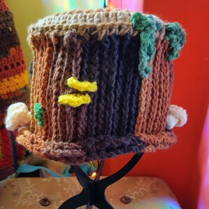 May include: A handmade crochet hat designed to resemble a tree stump. The hat features brown, tan, and dark blue yarn, with yellow mushroom accents and green leaves. The brim is a darker brown, and the top is a lighter tan.