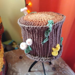 May include: A handmade, knitted tree stump-shaped container. The brown and tan top is detailed to resemble wood grain. The sides are a ribbed, dark brown knit, adorned with white flowers, green leaves, and a yellow mushroom. The container sits on a black, three-legged stand.