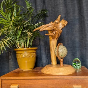 Könnte beinhalten: Eine Holzskulptur eines Baumzweigs mit einem Globus und einer grünen Glaskugel auf einem Holztisch mit einer Topfpflanze.