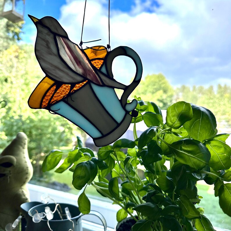 K&ouml;nnte beinhalten: Ein Glasfenster-Vogel sitzt auf einer blau-grauen Teetasse. Der Vogel hat graue und burgunderfarbene Fl&uuml;gel, und die Tasse hat einen gelb-orangenen Rand. Die Tasse h&auml;ngt vor einem Fenster mit einer Basilikumpflanze.