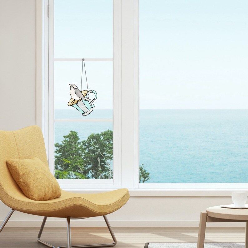 K&ouml;nnte beinhalten: Ein Fensterbehang aus Buntglas mit einem Vogel auf einer Teetasse. Die Teetasse ist hellblau mit gelben und wei&szlig;en Details. Der Vogel ist grau und wei&szlig;. Der Fensterbehang h&auml;ngt vor einem Fenster mit Blick auf das Meer.