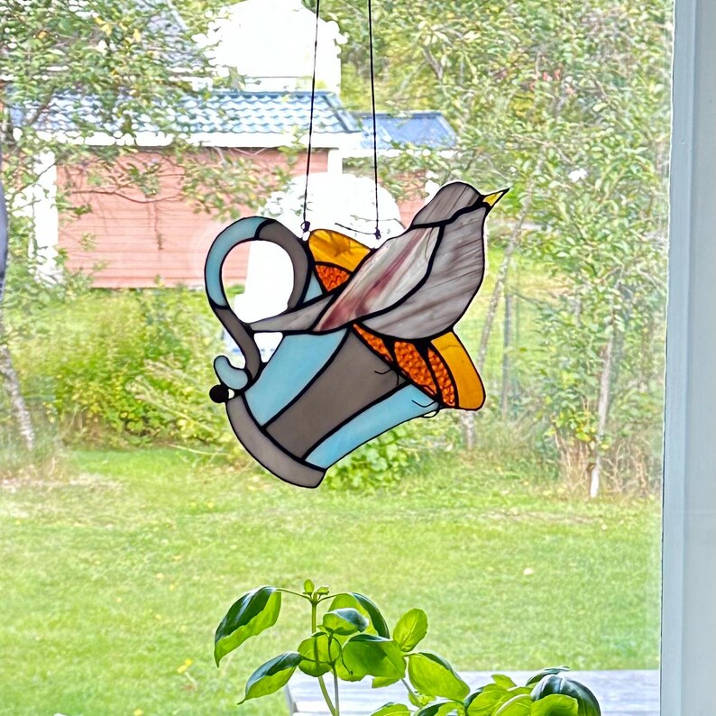 K&ouml;nnte beinhalten: Buntglas-Kunstwerk eines Vogels auf einer Teetasse. Die Teetasse ist blau, braun und orange. Der Vogel ist grau und braun mit einem gelben Schnabel. Das Kunstwerk wird von einer schwarzen Schnur gehalten.