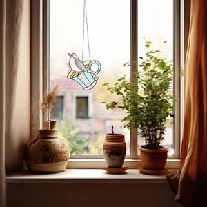 K&ouml;nnte beinhalten: Ein Vogelfutterhaus aus Buntglas in Form einer Teetasse, das vor einem Fenster h&auml;ngt. Das Futterhaus ist hellblau mit einem gelben Akzent und einem grauen Vogel, der darauf sitzt. Auf der Fensterbank befinden sich eine Vase, ein kleiner Topf und eine Topfpflanze.