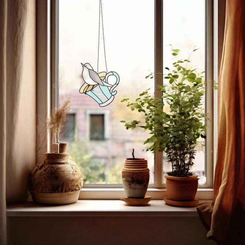 K&ouml;nnte beinhalten: Ein Vogelfutterhaus aus Buntglas in Form einer Teetasse, das vor einem Fenster h&auml;ngt. Das Futterhaus ist hellblau mit einem gelben Akzent und einem grauen Vogel, der darauf sitzt. Auf der Fensterbank befinden sich eine Vase, ein kleiner Topf und eine Topfpflanze.