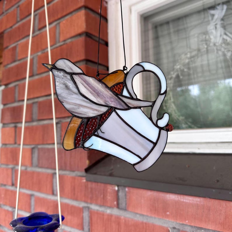 K&ouml;nnte beinhalten: Vogelhaus aus Buntglas mit einem Vogel, der auf einer dekorativen Tasse sitzt. Der Vogel hat graue und violette Fl&uuml;gel, und die Tasse ist wei&szlig; und grau mit einem Henkel. Das Vogelhaus h&auml;ngt vor einer Backsteinmauer und einem Fenster.