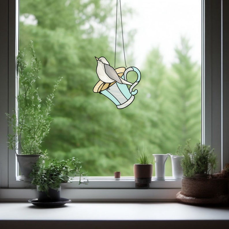 K&ouml;nnte beinhalten: Ein Fensterbehang aus Buntglas mit einem Vogel auf einer Teetasse. Die Teetasse ist hellblau mit wei&szlig;en und goldenen Akzenten. Der Vogel ist grau und wei&szlig;. Das Fenster h&auml;ngt an einer Kette vor einem Fenster mit gr&uuml;nem Hintergrund.