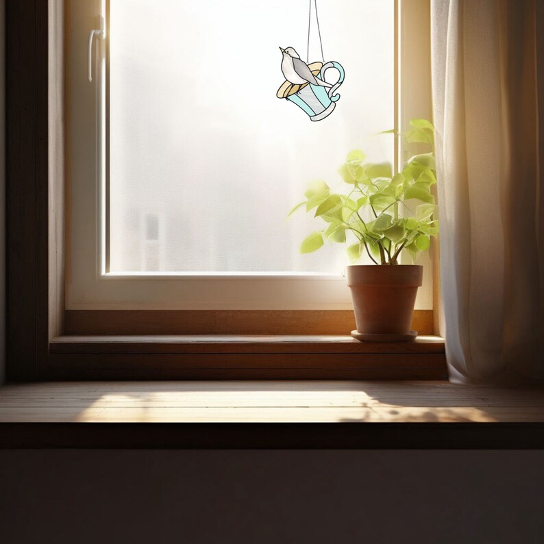 K&ouml;nnte beinhalten: Ein Glasfenster-Sonnenf&auml;nger mit einem Vogel auf einer Teetasse. Die Teetasse ist hellblau mit gelben und wei&szlig;en Akzenten. Der Vogel ist grau und wei&szlig;. Der Sonnenf&auml;nger h&auml;ngt vor einem Fenster mit einer Topfpflanze auf der Fensterbank.