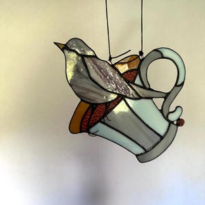 K&ouml;nnte beinhalten: Glasmalerei Vogel sitzt auf einer hellblauen und grauen dekorativen Kanne. Der Vogel hat graue und violette Fl&uuml;gel und einen gelben Schnabel. Die Kanne hat ein rotes und orangefarbenes Design. Schwarze Schnur zum Aufh&auml;ngen.