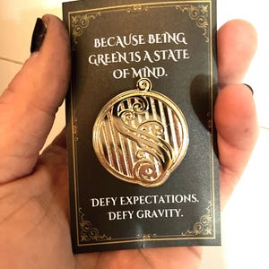 May include: Gold-colored pin with a striped design and a swirl. The pin is on a black card with gold lettering that reads "Because being green is a state of mind. Defy expectations. Defy gravity."