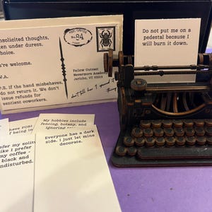 K&ouml;nnte beinhalten: Eine Miniatur-Schreibmaschine im Vintage-Stil aus dunklem Holz, mit einem kleinen Papierst&uuml;ck im Wagen. Daneben liegen mehrere Karten mit Text, darunter eine Postkarte mit einer Briefmarke und dem Text "Meine unaufgeforderten Gedanken."