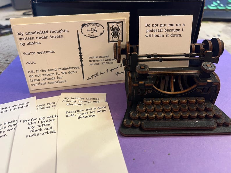 K&ouml;nnte beinhalten: Eine Miniatur-Schreibmaschine im Vintage-Stil aus dunklem Holz, mit einem kleinen Papierst&uuml;ck im Wagen. Daneben liegen mehrere Karten mit Text, darunter eine Postkarte mit einer Briefmarke und dem Text "Meine unaufgeforderten Gedanken."