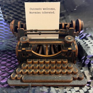 K&ouml;nnte beinhalten: Eine Miniatur-Schreibmaschine in Schwarz und Braun mit einem Papier mit der Aufschrift "Outcasts welcome, Normies tolerated." Die Schreibmaschine hat ein detailliertes Design mit Tastatur und Walzen und ist aus Holz gefertigt.